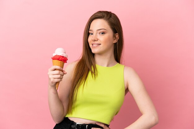 Menina adolescente com um sorvete de corneta sobre um fundo rosa isolado, posando com os braços na cintura e sorrindo