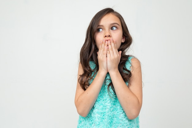 Foto menina adolescente com um vestido azul cobriu a boca com as mãos em um fundo branco do estúdio.