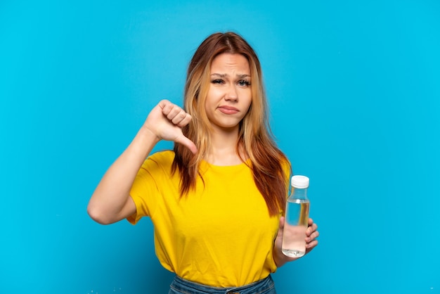 Menina adolescente com uma garrafa de água sobre um fundo azul isolado, mostrando o polegar para baixo com expressão negativa