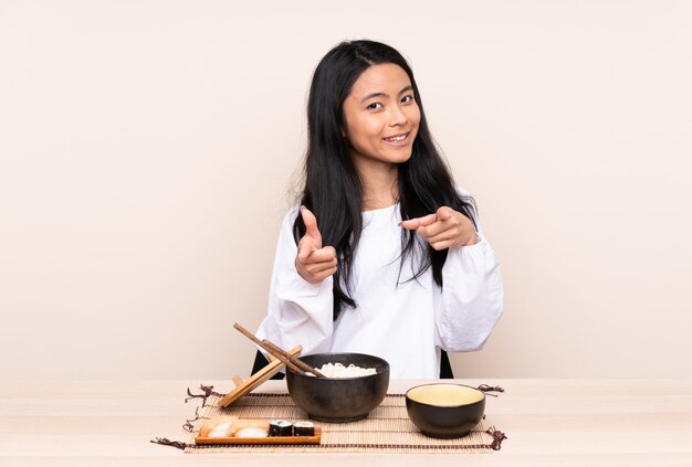 Menina adolescente comendo comida asiática isolada em bege, apontando para a frente e sorrindo