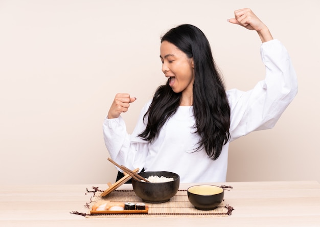 Menina adolescente comendo comida asiática isolada em bege comemorando uma vitória