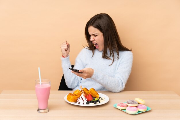 Menina adolescente comendo waffles isolados na parede bege com telefone em posição de vitória