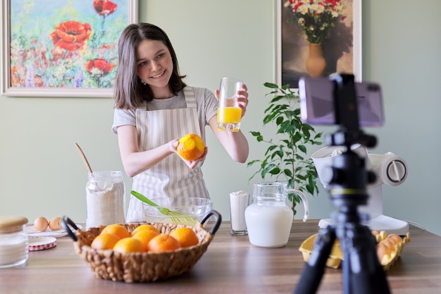 Menina adolescente comida blogger gravando receita para cozinhar panquecas de laranja. mulher em casa na cozinha, espreme suco, sorri e olha para a câmera, hobby e lazer, conceito de comida caseira saborosa saudável