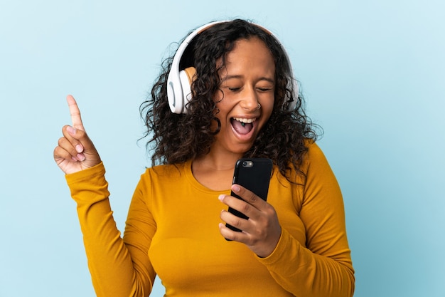 Menina adolescente cubana isolada em um fundo azul, ouvindo música com um celular e cantando