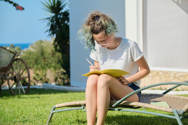 Menina adolescente de 15, 16 anos sentada numa espreguiçadeira no gramado, escrevendo com o lápis no caderno. De volta às aulas, início das aulas no verão