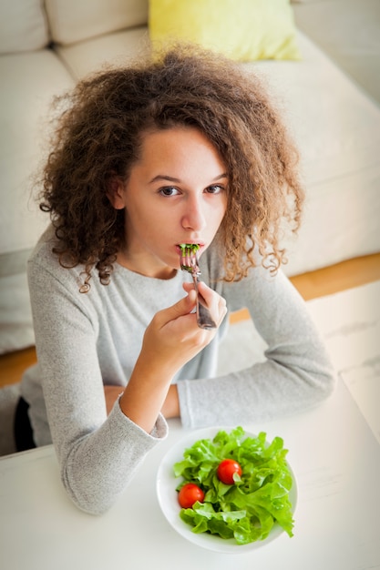 Menina adolescente de cabelo crespo comendo salada saudável no quarto