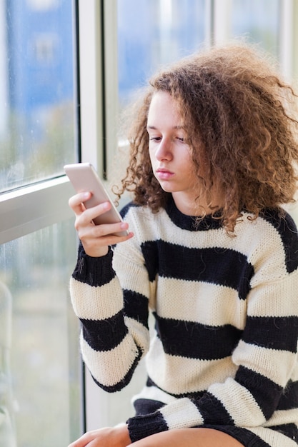 Menina adolescente de cabelo encaracolado com telefone móvel pela janela