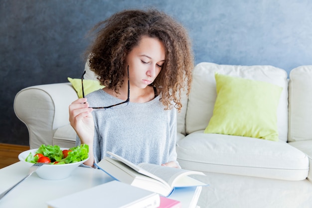 Menina adolescente de cabelo encaracolado lendo livro e comendo salada