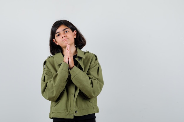 Menina adolescente de mãos dadas em gesto de oração em t-shirt, jaqueta verde e olhando esperançosa. vista frontal.