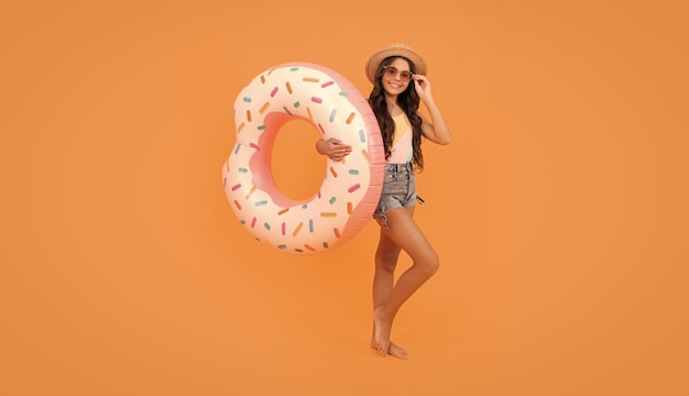 Menina adolescente de praia feliz com cabelo encaracolado com chapéu de palha e óculos de sol com anel inflável de rosquinha para diversão de festa na piscina nas férias de verão no acessório de praia de fundo laranja