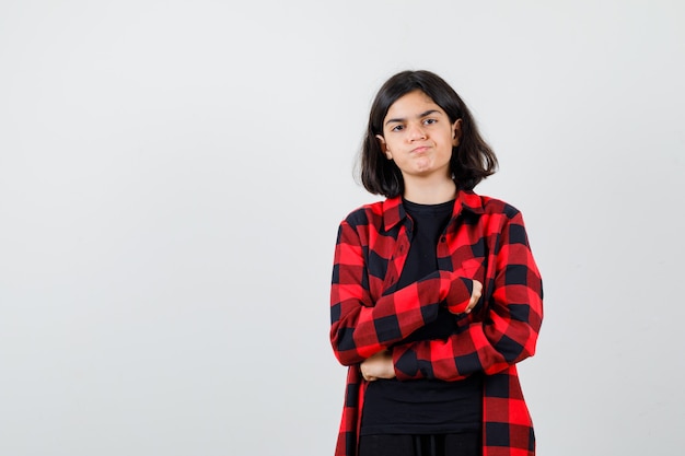 Menina adolescente em pé com os braços cruzados em t-shirt, camisa xadrez e olhando imprudente, vista frontal.