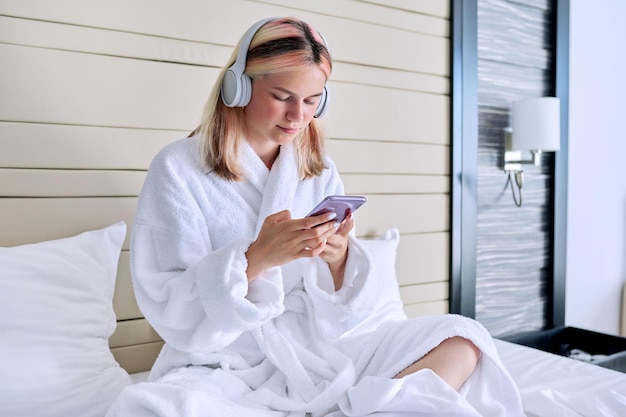 Menina adolescente em roupão branco e fones de ouvido com smartphone nas mãos