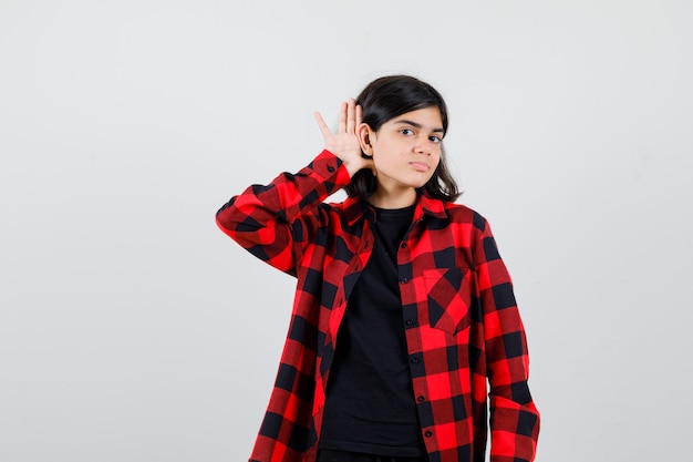 Menina adolescente em t-shirt, camisa quadriculada, segurando a mão atrás da orelha e olhando com foco, vista frontal.