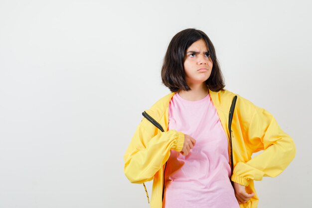 Menina adolescente em t-shirt, jaqueta, apontando para si mesma e parecendo confusa, vista frontal.