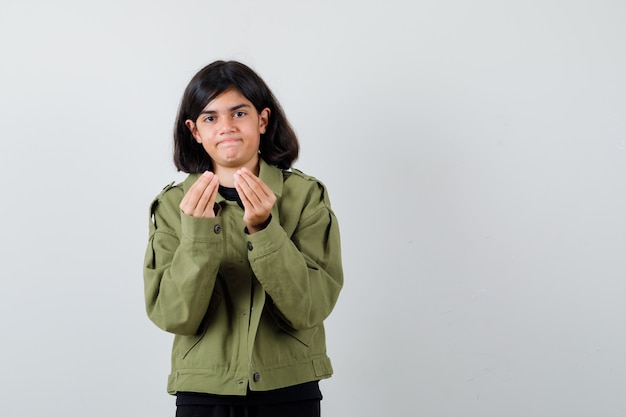 Menina adolescente em t-shirt, jaqueta verde mostrando gesto italiano e parecendo insatisfeita, vista frontal.