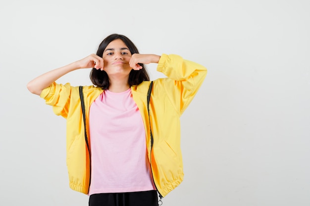 Menina adolescente em um agasalho amarelo, camiseta puxando as bochechas para os lados e parecendo melancólica, vista frontal.