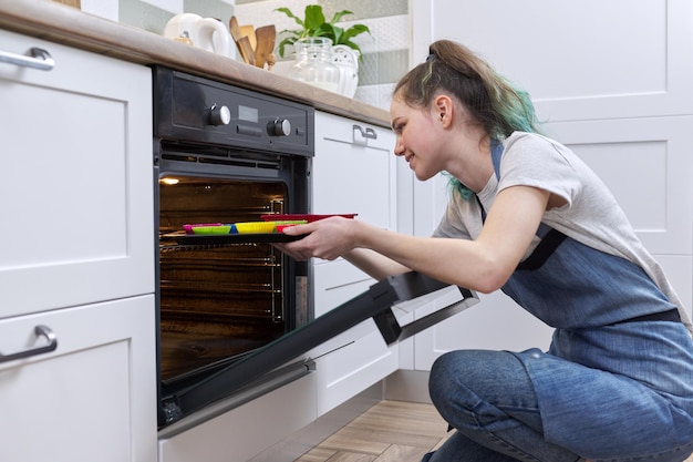 Menina adolescente em um avental com a preparação de cupcakes crus em moldes de silicone, colocando a bandeja no forno, interior de plano de fundo de comida caseira. Hobbies adolescentes, cozinhar em casa, comida caseira saudável