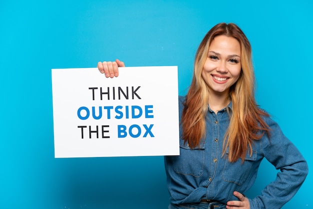 Menina adolescente em um fundo azul isolado segurando um cartaz com o texto pense fora da caixa com uma expressão feliz