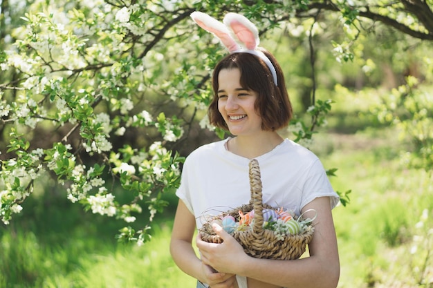 Menina adolescente engraçada com cesta de ovos e orelhas de coelho na caça aos ovos de páscoa no jardim ensolarado da primavera