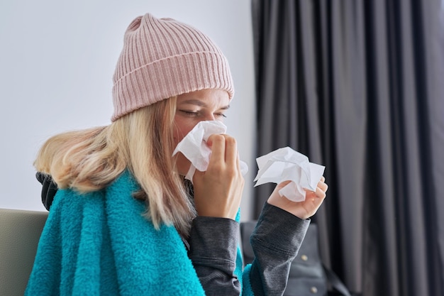 Menina adolescente espirrando no lenço, fêmea com sintomas de doença sentada em casa coberta com cobertor quente e chapéu de malha