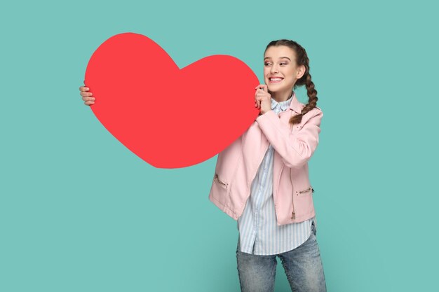 Foto menina adolescente extremamente feliz com tranças mostrando um grande coração vermelho olhando para longe com um sorriso