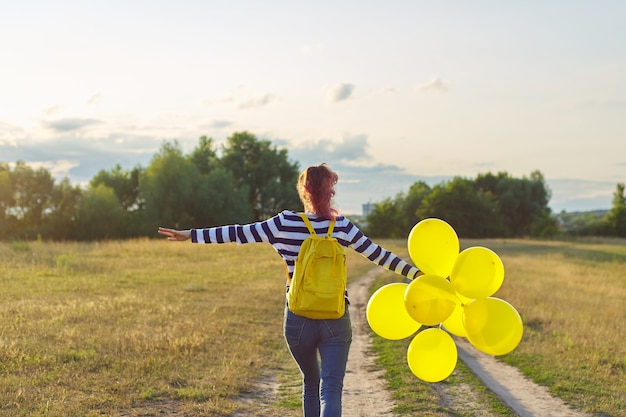 Menina adolescente feliz com balões amarelos e mochila correndo e pulando ao longo da estrada rural num prado de verão. Liberdade, vida, alegria, conceito de férias
