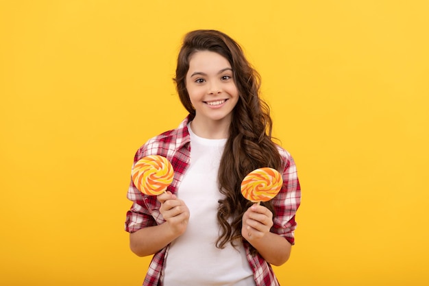 menina adolescente feliz com cabelo longo encaracolado em camisa quadriculada segura pirulito caramelo doce em fundo amarelo, loja de caramelo.