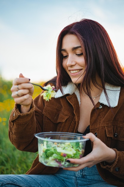 Menina adolescente feliz comendo uma salada enquanto está sentado na grama em uma colina Ela está aproveitando a calma