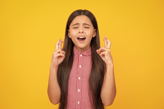 Menina adolescente feliz cruza os dedos fecha os olhos com prazer antecipar boas notícias isolado fundo amarelo Criança feliz se alegra que seus sonhos se tornaram realidade Conceito de excitação