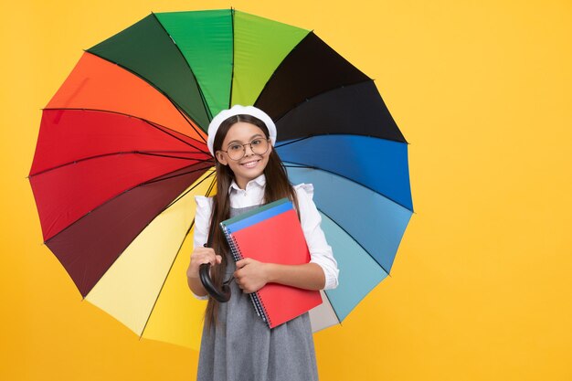 Menina adolescente feliz de óculos e boina sob guarda-chuva colorida para proteção contra chuva na temporada de outono segura o notebook de volta à escola