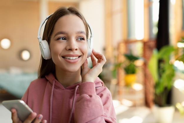 Menina adolescente feliz em fones de ouvido usando telefone celular curtindo música ou ouvindo livro de áudio sentado em