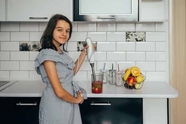 Menina adolescente feliz fazendo suco fresco ou refeição no liquidificador na cozinha.