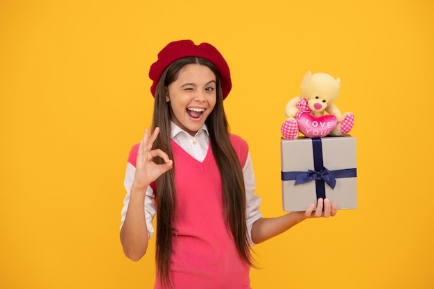 Menina adolescente feliz na boina francesa segura o brinquedo presente e a caixa de presente mostra o gesto okey presente