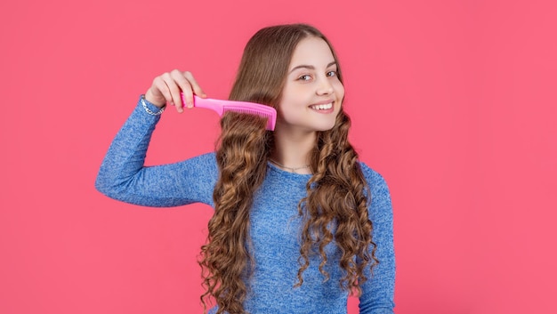 Menina adolescente feliz penteando o cabelo encaracolado com escova de cabelo no fundo rosa