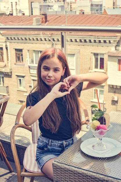 Foto menina adolescente feliz sorrindo, fazendo um gesto de coração com as mãos, sentado no restaurante ou café e comendo sorvete. comer