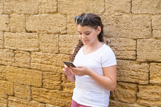 Menina adolescente feliz verificando mídias sociais segurando smartphone n cidade sobre fundo de parede de pedra