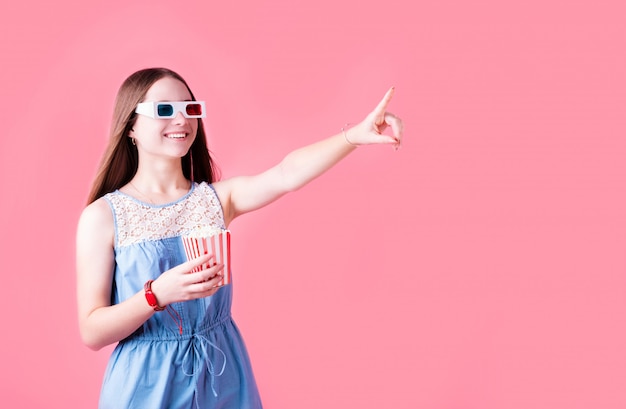 Menina adolescente feminino usando óculos 3D, comendo pipoca e apontando o dedo isolado no fundo rosa