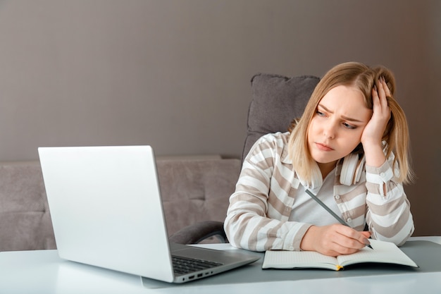 Menina adolescente infeliz durante a educação on-line em casa usando o laptop. Aluna com tristeza de estresse faz aulas de dever de casa. Mulher triste trabalhar longe de casa.