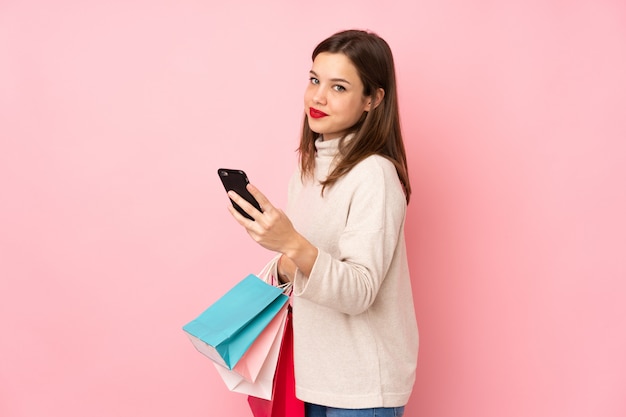 Menina adolescente isolada na parede rosa segurando sacolas de compras e escrever uma mensagem com o celular para um amigo