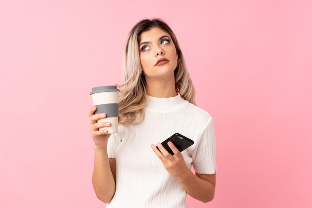 Menina adolescente isolado rosa segurando café para levar embora e um móvel enquanto pensava em algo