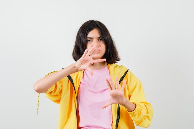 Menina adolescente mostrando gesto de parada em agasalho amarelo, camiseta e olhando melancólica, vista frontal.