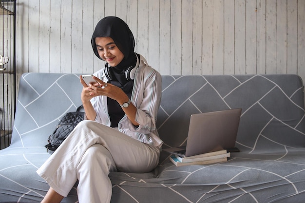 Menina adolescente muçulmana asiática verificando as mídias sociais segurando o smartphone em casa. usando o jogo do aplicativo do telefone móvel