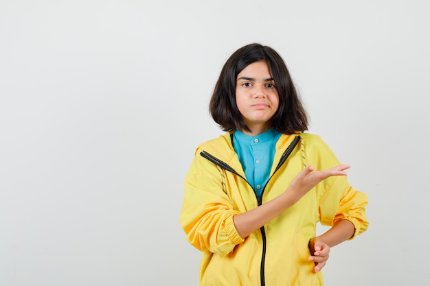 Menina adolescente na jaqueta amarela fingindo mostrar algo e parecendo descontente, vista frontal.