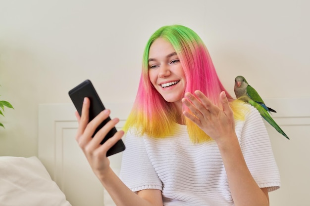 Menina adolescente na moda com smartphone e papagaio quaker verde jovem no ombro, em casa na cama. Proprietário do animal de estimação falando em comunicação de vídeo, amizade de pássaros humanos, estilo de vida, comunicação