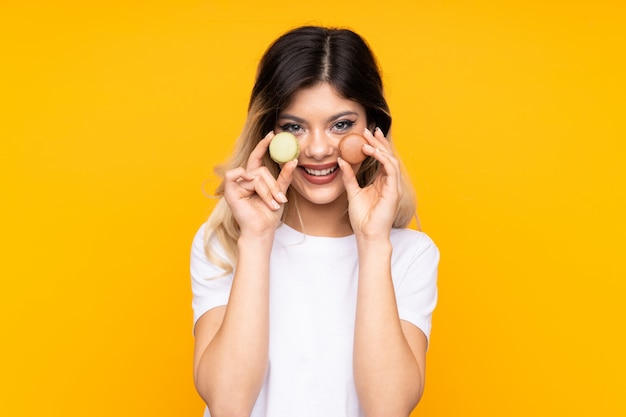 Menina adolescente na parede amarela segurando macarons franceses coloridos e sorrindo