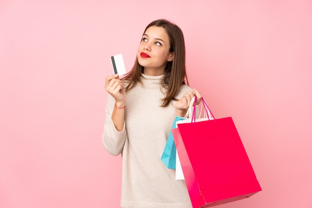 Menina adolescente na parede rosa segurando sacolas de compras e um cartão de crédito e pensando