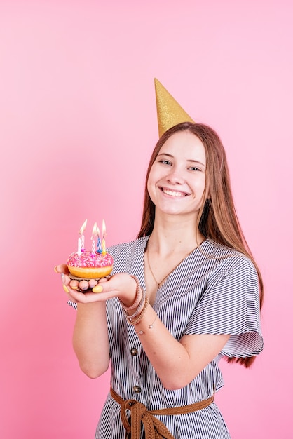 Menina adolescente no aniversário de ouro segurando um donut com uma vela e fazendo um pedido sobre o fundo rosa