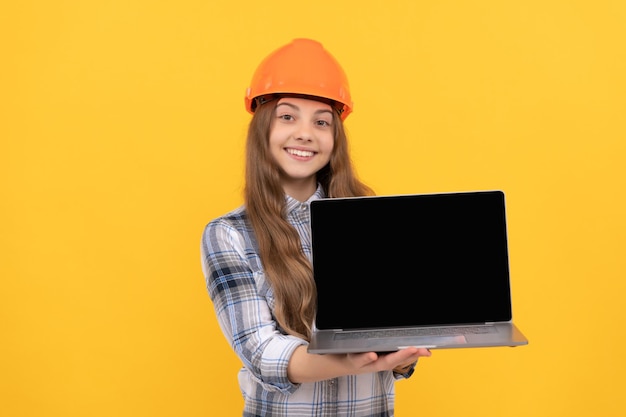 menina adolescente no capacete apresentando laptop. garoto construtor segura computador. construção e construção. apresentação. trabalhador criança feliz usa capacete. educação online infantil. feliz Dia do Trabalho. copie o espaço.