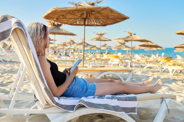 Menina adolescente olhando para a tela do smartphone sentado na espreguiçadeira fim de semana na praia