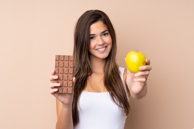 Menina adolescente parede isolada tomando uma tablete de chocolate em uma mão e uma maçã na outra